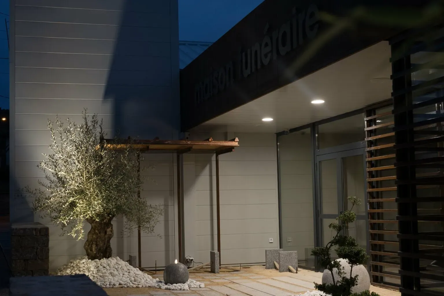 Entrée éclairée de la maison funéraire de nuit avec un olivier décoratif.